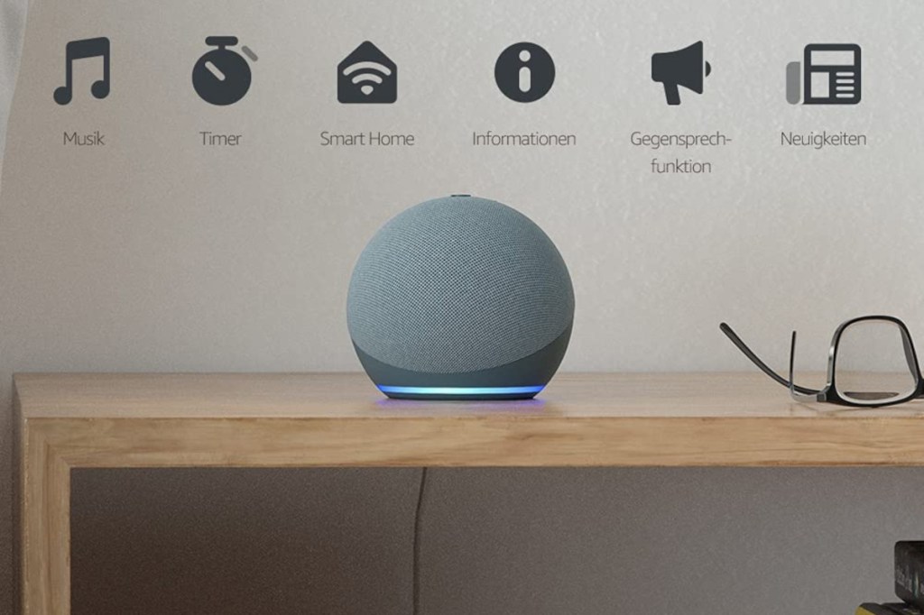 Grauer Amazon Echo Dot auf Holztisch auf dem eine Brille liegt vor grauer Wand, oben Icons der Funktionen