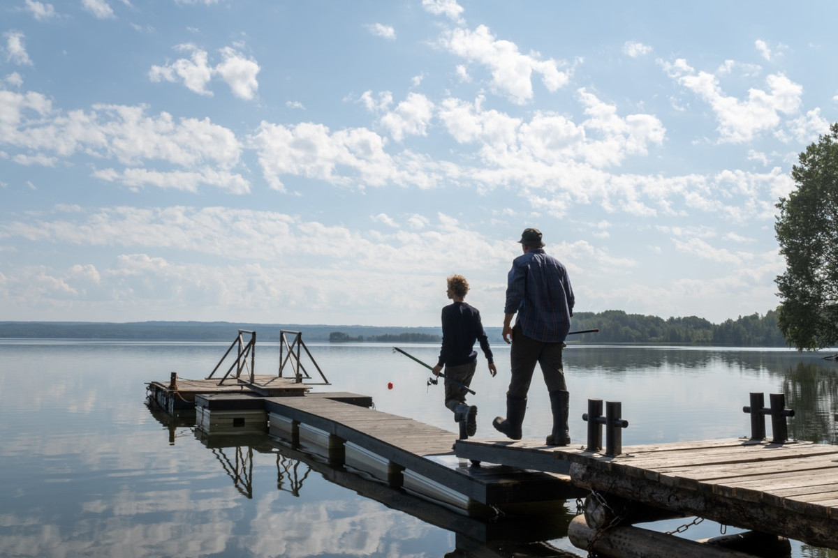 Mann und Junge in Anglerkluft gehen Steg an weiten See entlang mit blauen Himmel