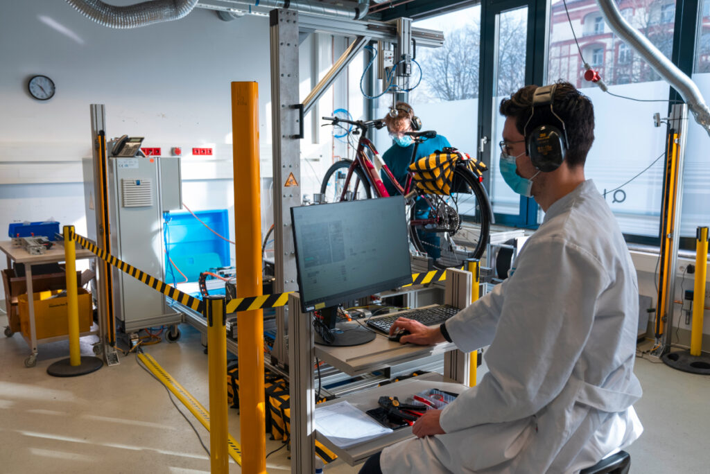 Mann in weißem Kittel sitzt vor Bildschirm, im Hintergrund Mann bei rotem E-Bike auf Prüfstand