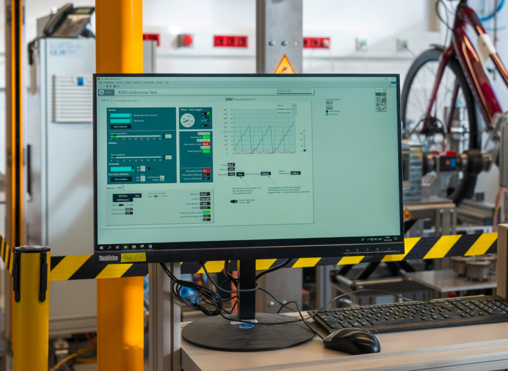 Monitor der Auswertungen anzeigt, im Hintergrund Vorderrad von rotem E-Bike auf Prüfstand