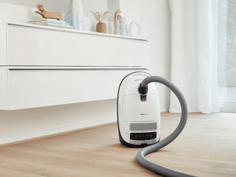 Weißer Miele-Staubsauger auf hellem Holzboden vor weißem Board