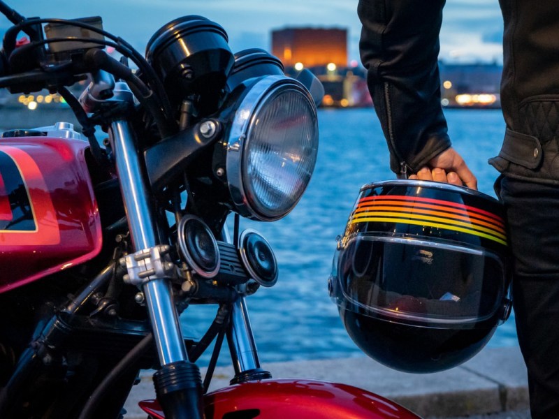 Rotes Motorrad Lenker im Bild, daneben mit Arm und Rücken von Mann, der bunten Motorradhelm hält