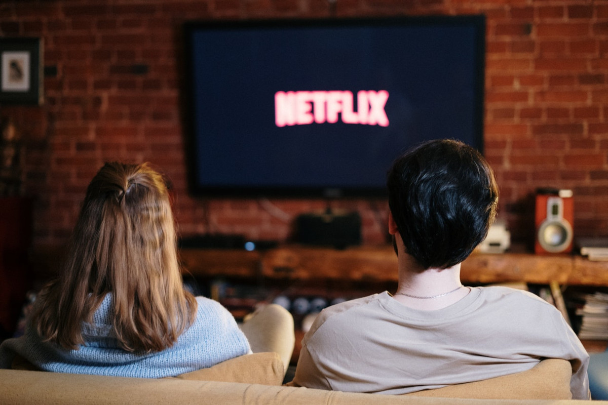 Frau und Mann sitzen auf Sofa von hinten vor großen Fernseher an Wand, der verschwommen Netflix Logo zeigt