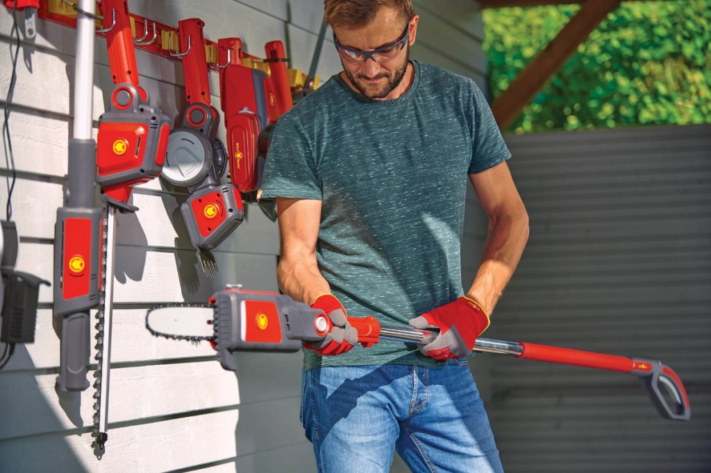 Mann in Jeans und blaugrauem T-Shirt mit Schutzbrille auf hantiert mit rotem Wolf e-Multi-Star Gerät vor weißer Holzwand an der weitere rote Gartenwerkzeuge hängen