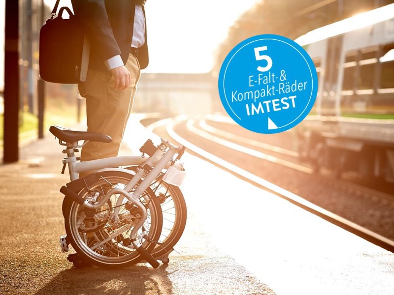 Ein Mann steht mit Falt-E-Bike auf Bahnsteig, von der Seite mit blauem Sticker "5 Kompakt- und Falt-E-Bikes IMTEST"