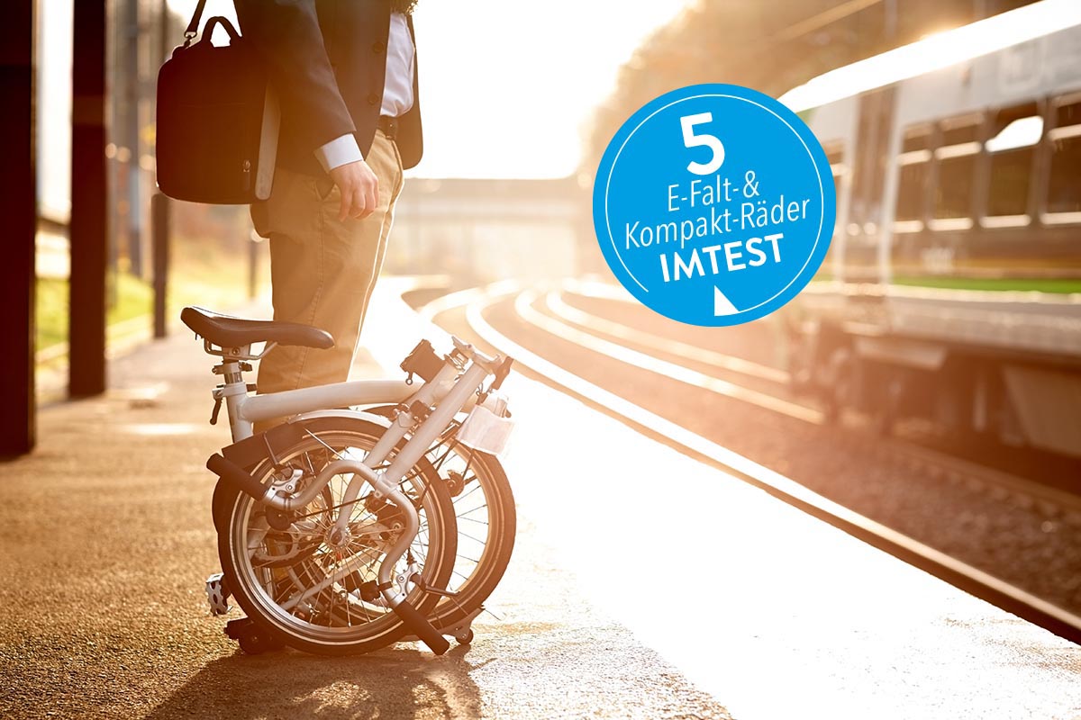 Ein Mann steht mit Falt-E-Bike auf Bahnsteig, von der Seite mit blauem Sticker "5 Kompakt- und Falt-E-Bikes IMTEST"