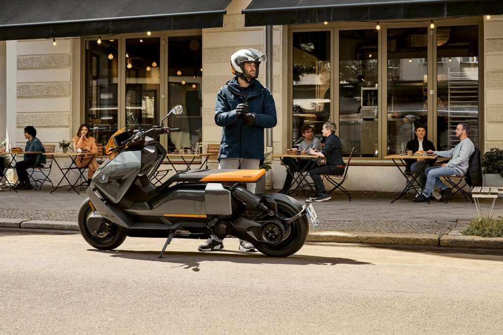 Mann mit Motorradhelm neben E-Roller, der vor einem Geschäft abgestellt ist.