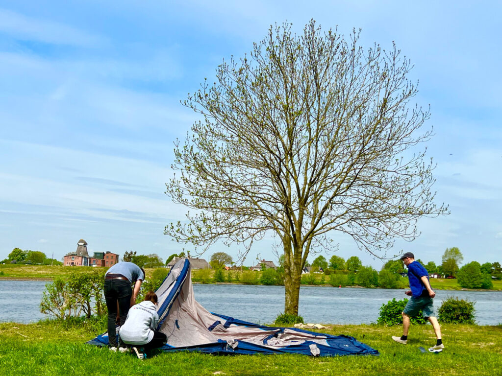 Auf Wiese an Flussufer bauen drei Personen Zelt auf