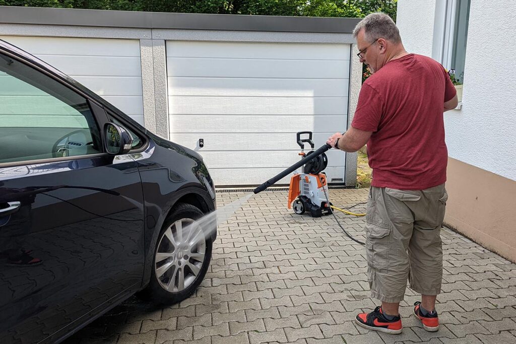 Mann in rotem Shirt mit Kaki-Hose hält Hochdruckreiniger auf Autoreifen