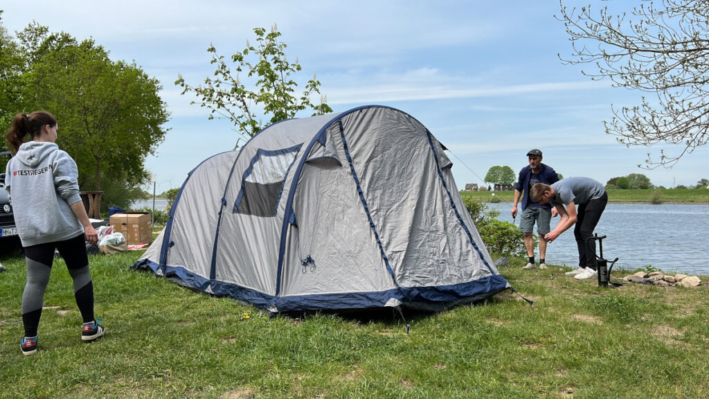 Helles Familienzelt wird auf Wiese von drei Personen aufgebaut