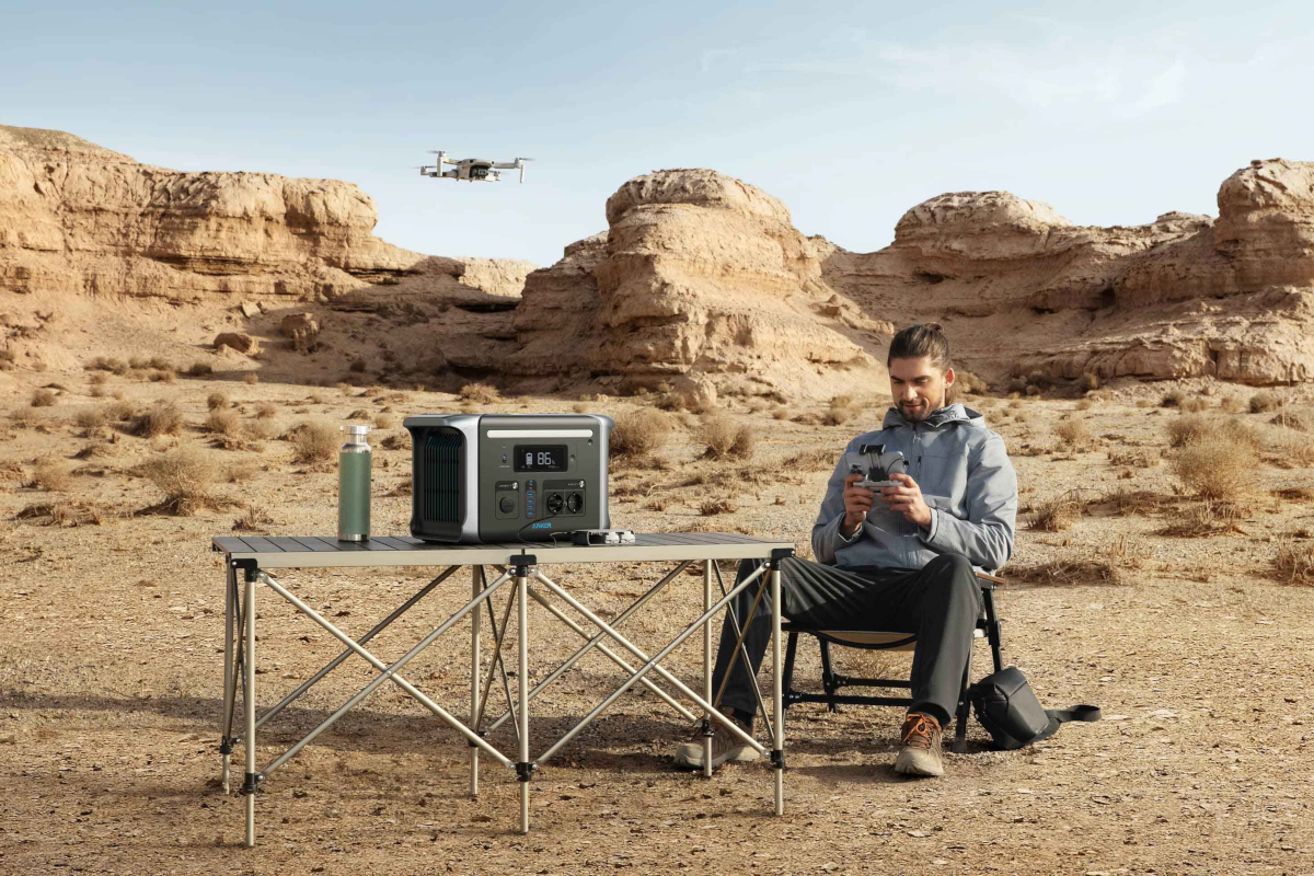 Mann grau gekleidet sitzt auf CampingStuhl neben Campingtisch auf dem die schwarze Anker PowerHouse steht, in Wüstenlandschaft mit fliegender Drohne im Hintergrund