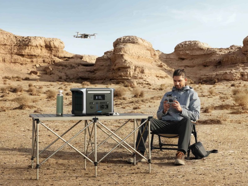 Mann grau gekleidet sitzt auf CampingStuhl neben Campingtisch auf dem die schwarze Anker PowerHouse steht, in Wüstenlandschaft mit fliegender Drohne im Hintergrund