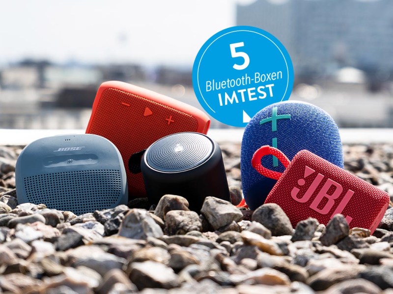 5 bunte Bluetooth-Boxen auf Kieselsteinen in Reihe angeordnet von vorne