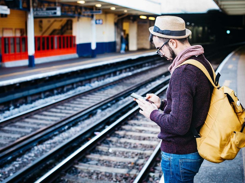 Ein Mann steht am Bahngleis und schaut auf sein Smartphone.