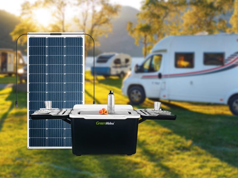 Es ist die GreenAkku Kühlbox im aufgeklappten Zustand sowie ein GreenAkku Solarmodul zu sehen. Im Hintergrund ist ein Campingplatz mit Wohnmobilen zu sehen.