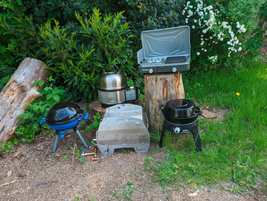 Fünf verschiedene mobile Gasgrills auf Wiese vor Büschen aufgestellt