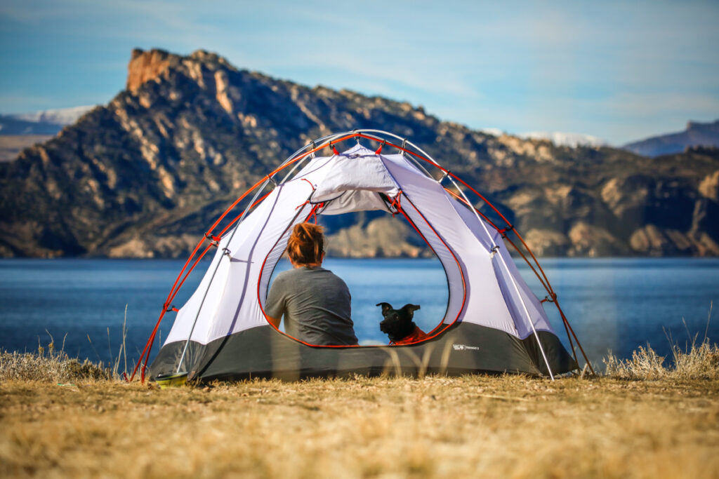 Eine Frau und ihr Hund sitzen in einem offenem Zelt vor einem See mit Bergen.