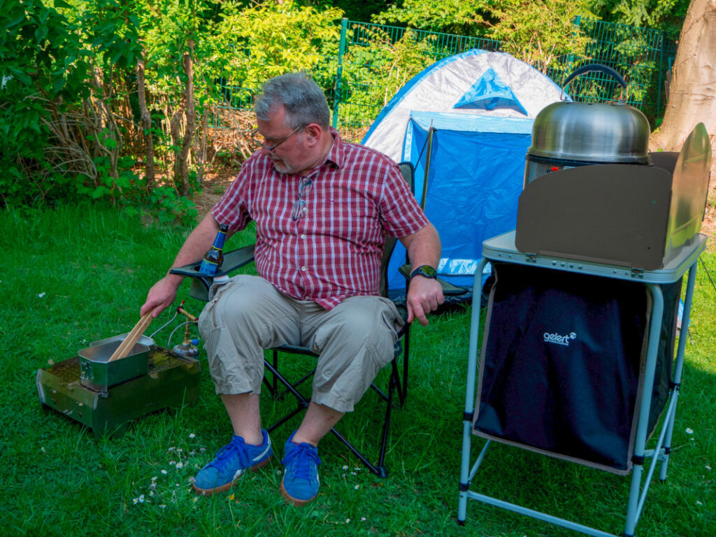 Mann sitzt in Campingstuhl zwischen Gasgrill und Camping-Küche vor blauem Zelt