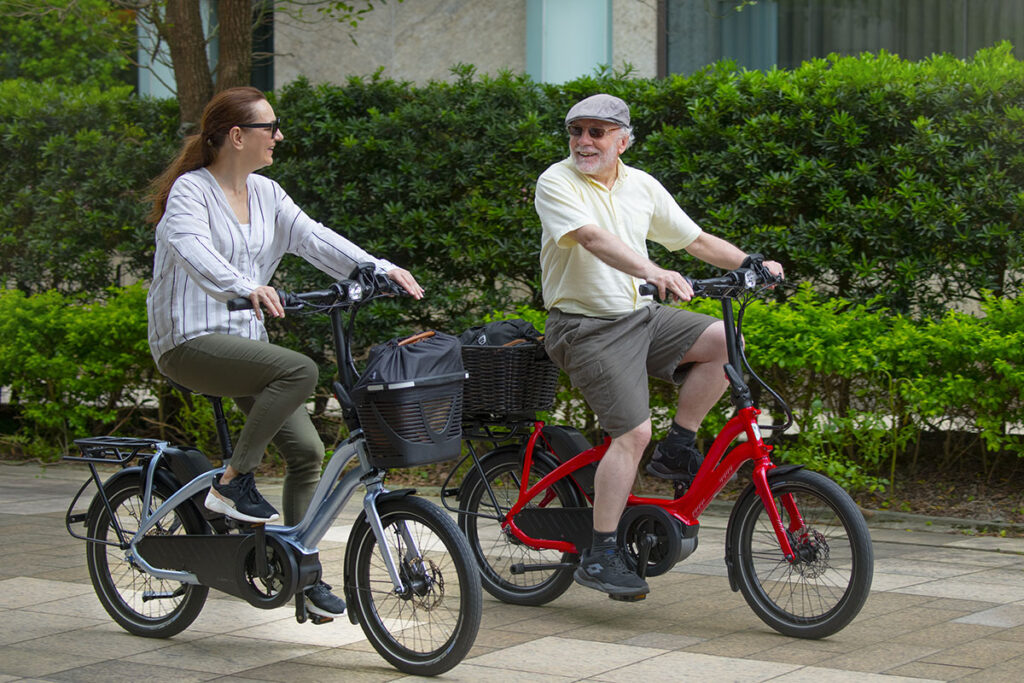 Eine ältere Frau und ein älterer Mann fahren je auf einem Tern NBD Kompakt-E-Bike an einer grüner Hecke vorbei.