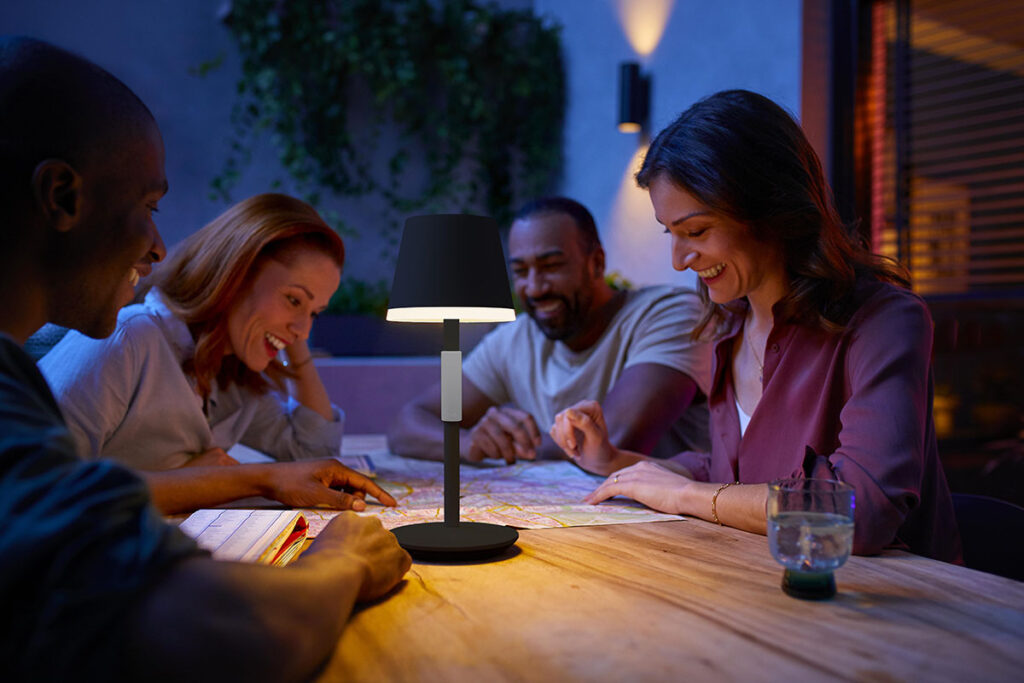 Zwei Frauen und ein Mann sitzen an einem Tisch und spielen ein Spiel, eine smarte Tischlanpe von Philips Hue  beleuchtet die Szenerie 