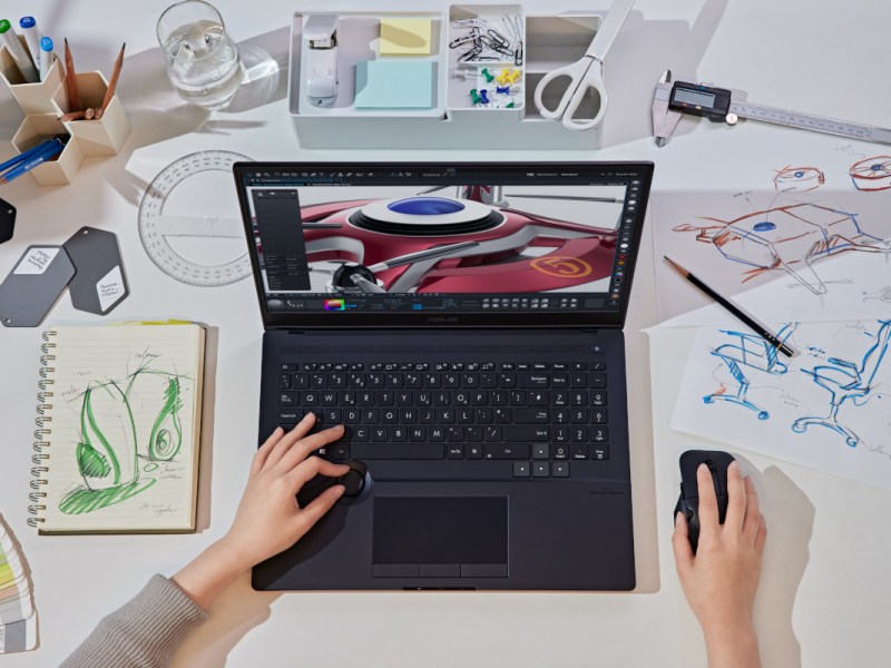 Asus ProArt Studiobook in schwarz aufgeklappt schräg von oben wird von zwei Händen bedient auf weißem Schreibtisch mit bunten Zeichnungen