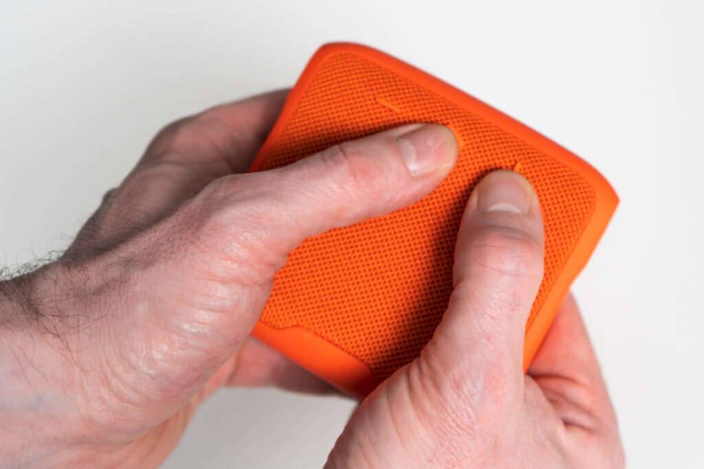 Zwei Hände halten orangene rechteckige Bluetooth-Box vor weißem Hintergrund