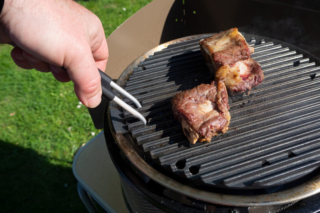 Hand hält Grillzange an Fleischstücke auf schwarzem Grill