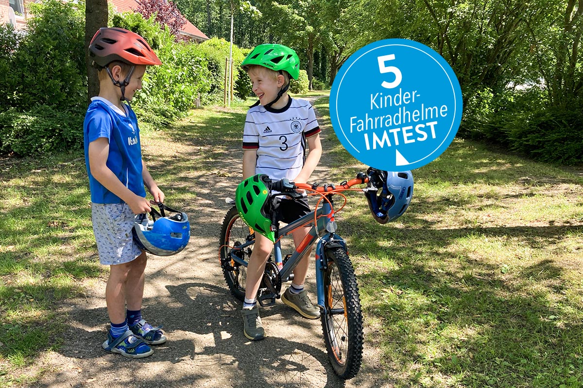 Zwei Kinder lachen sich an, das eine sitzt auf einem Fahrrad und trägt ein Helm, zwei weitere Helme sind am Lenker gefestigt, das zweite Kind steht daneben. Es hat ebenfalls einen Helm auf und hält zudem einen in der Hand