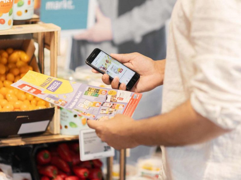 Eine Person hält einen Werbeprospekt von Rewe in der Hand sowie die Rewe App auf dem Handy.