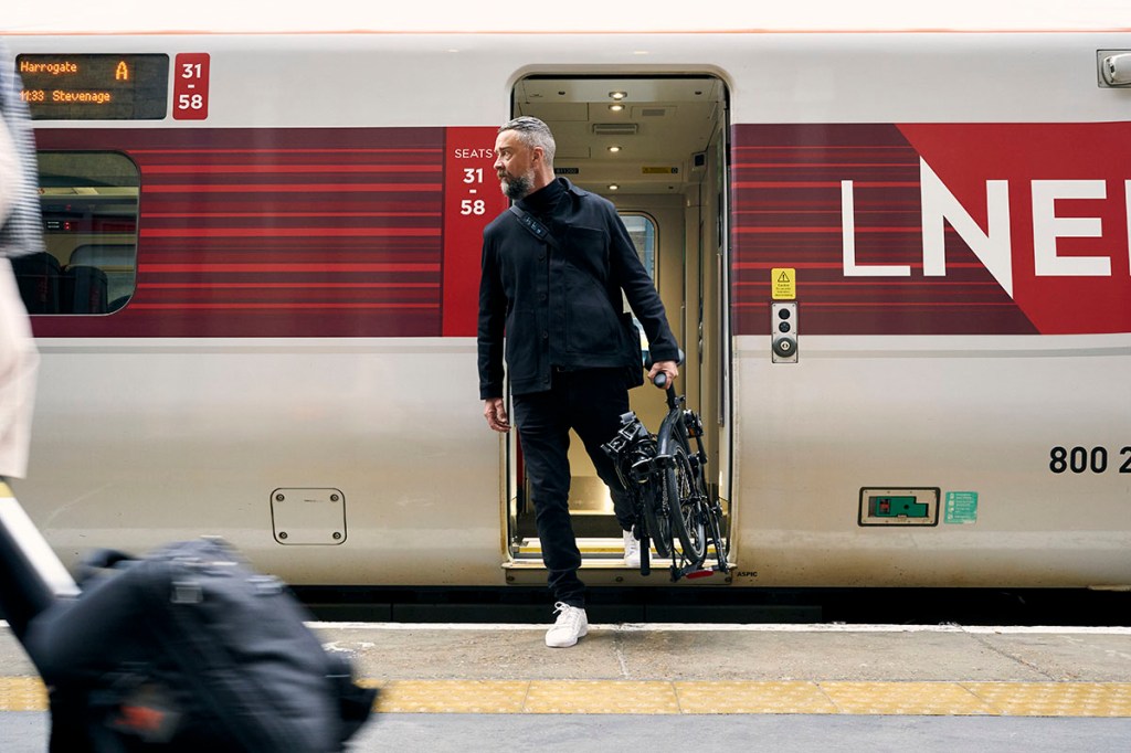 Brompton Electric P Line: Mann trägt Faltbares E-Bike mit einer Hand aus einem Zugabteil in einen Bahnhof.