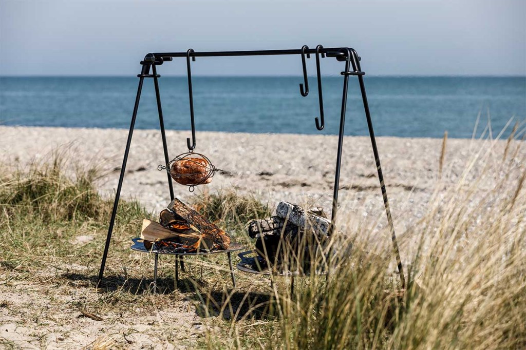 Der Petromax Geflügel-Korb hängt mit einem Hähnchen befüllt über einem Lagerfeuer am Strand.