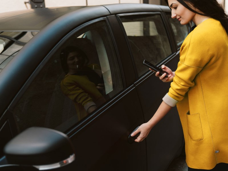 Frau mit Smartphone vor Solarauto. DasSolarauto Sion und normale PKW können über Sono-App miteiender getilet werden.