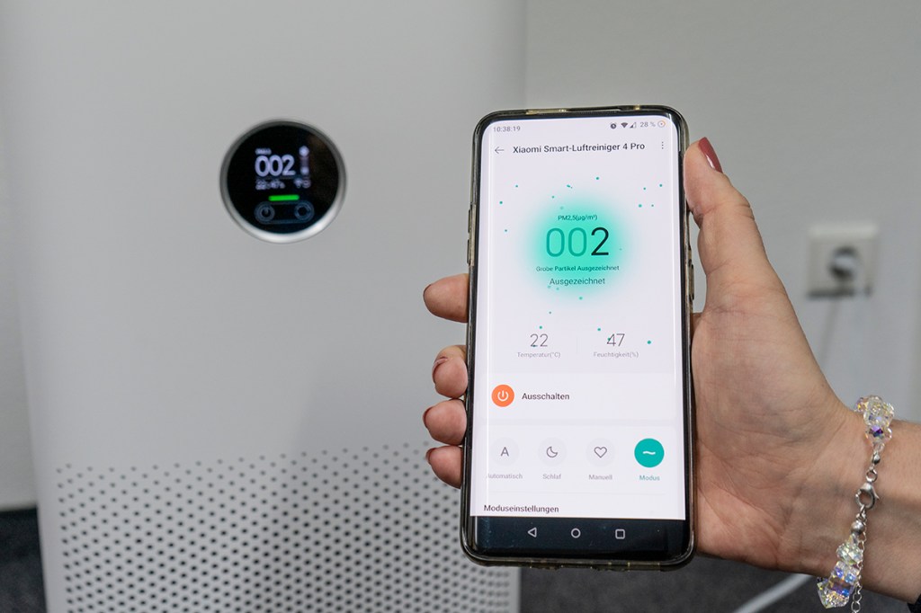Hand hält Smartphone ins Bild mit App die türkisen Kreis und Zahl anzeigt vor Anzeige von Luftreiniger, die das gleiche anzeigt