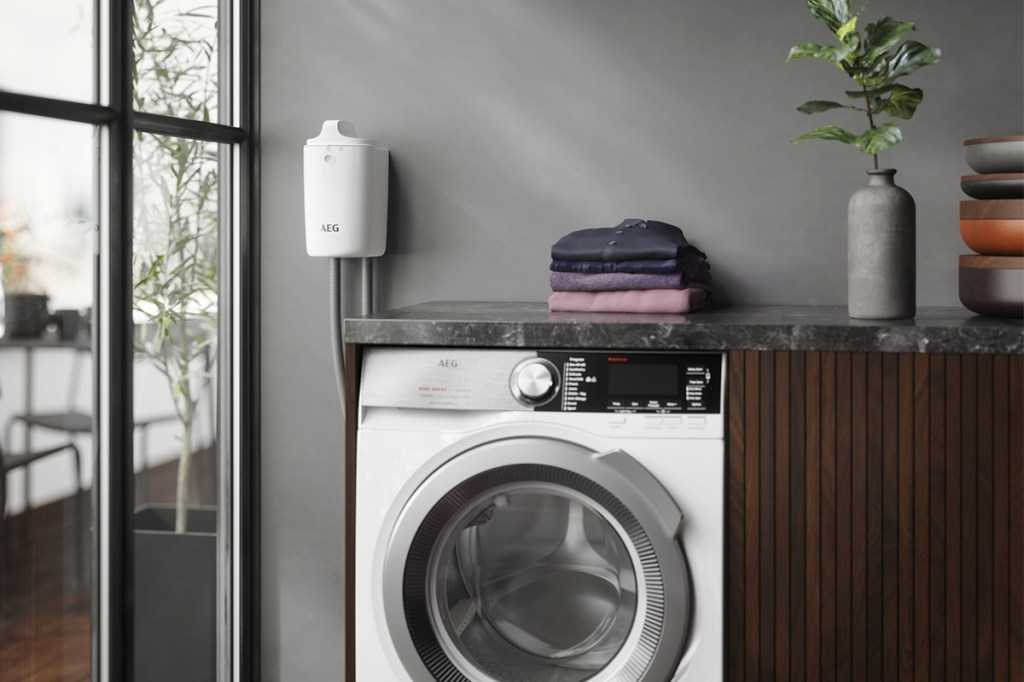 Das Bild zeigt eine AEG Waschmaschine vor einer grauen Wand. Links neben der Maschine ist der Mikroplastikfilter als weiße Box an der Wand angebracht.