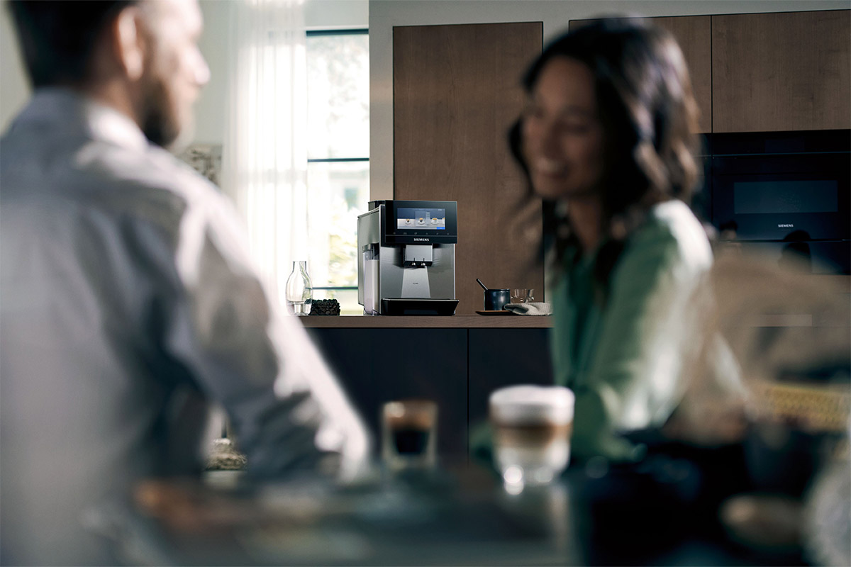 Im Vordergrund sind verschwommen zwei Personen zu sehen, die an einem Tisch sitzen und unterschiedliche Kaffee-Getränke trinken. Im Fokus steht der neue Siemens EQ900-Kaffeevollautomat.