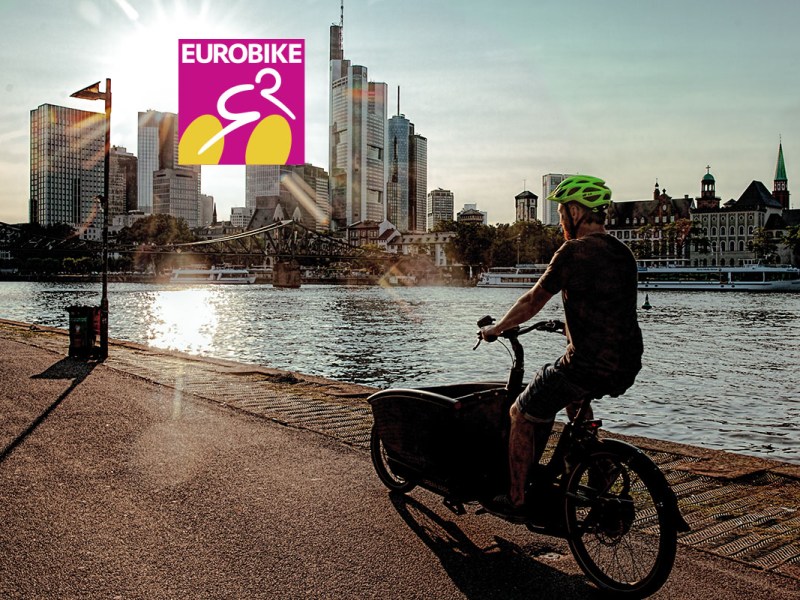 E-Bike-Markt weiter auf Rekordniveau: Mann fährt mit E-Cargobike an Fluss vor Stadtsilhouette entlang.