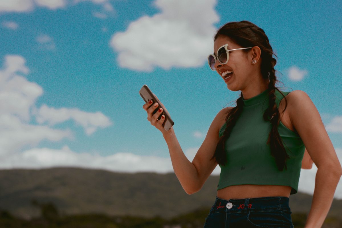 Eine Frau im grünen Tank Top mit weißer Sonnenbrille schaut lachend auf ihr Smartphone. Im Hintergrund ein heiterer Himmel und Berge.