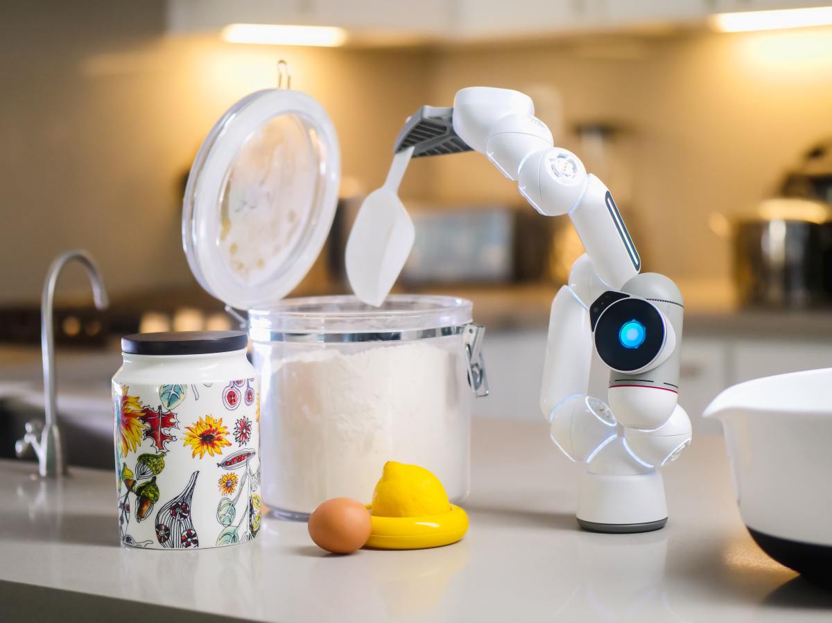 Es ist ein kleiner Roboter auf einer Arbeitsfläche zu sehen, der aus mit einem Löffel Mehl aus einem Gefäß entnimmt.