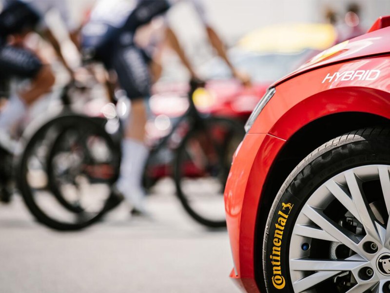 Angeschnittenes Auto mit Reifen im Vordergrund, im Hintergrund in der Unschärfe Fahrradfahrer