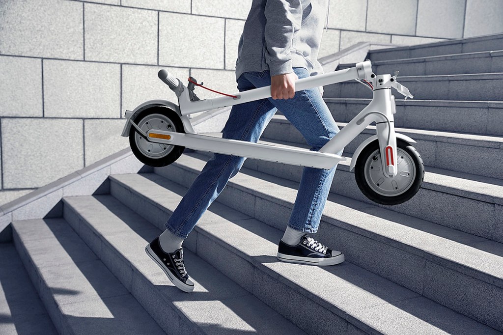 Eine Frau trägt einen zusammengeklappten E-Scoooter eine Treppe hinauf.