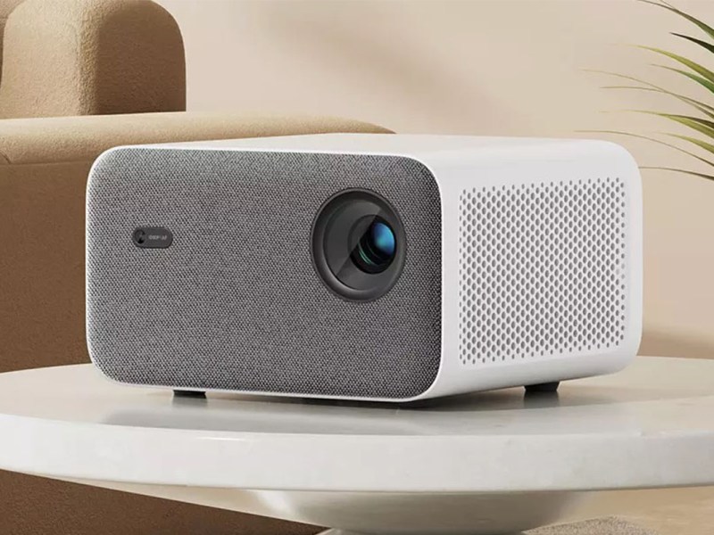 Der Mi Projector 2S ist als Nahaufnahme zu sehen. Er steht auf einem Beistelltisch in einem Zimmer mit Sofa und Topfpflanze.