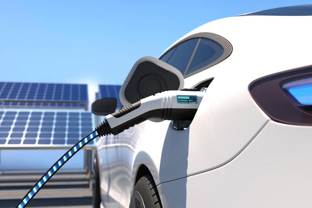 Weißes E-Auto wird vor Solarpanels mit einem Stecker geladen