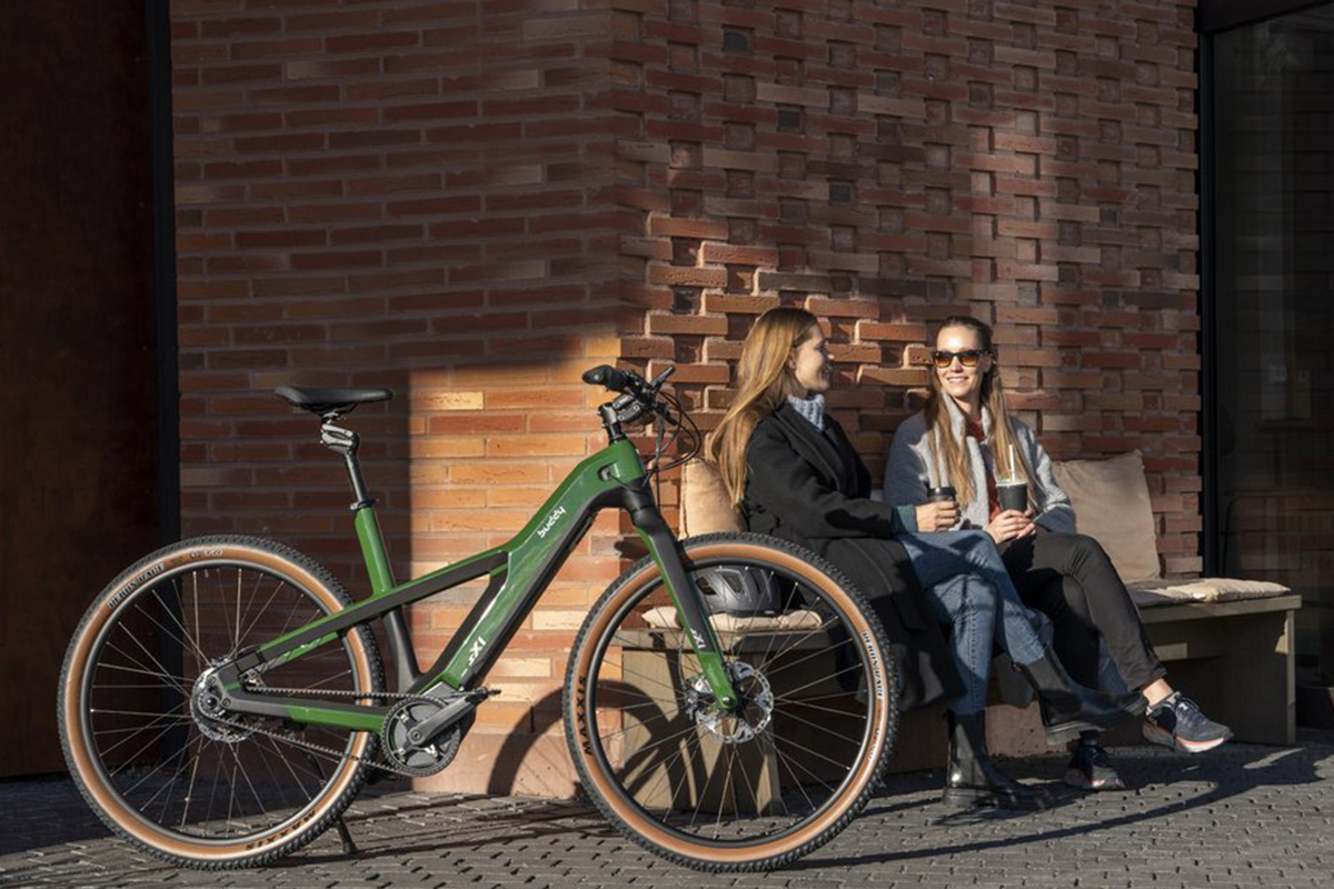 E-bike sX1 von Buddy Bike an einer Mauer gelehnt, dahinter sitzen auf einer Bank zwei Frauen