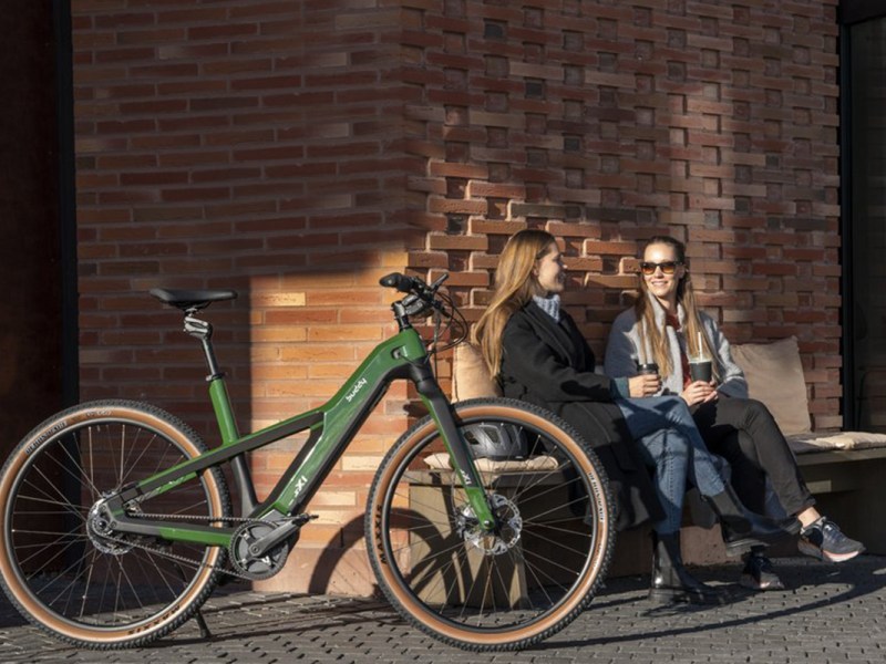 E-bike sX1 von Buddy Bike an einer Mauer gelehnt, dahinter sitzen auf einer Bank zwei Frauen