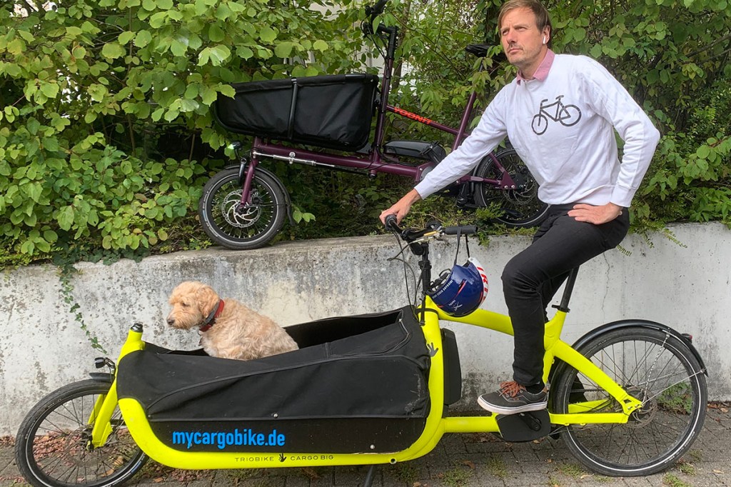 E-Cargo-Bike-Fahrer steht mit seinem Rad an einer Mauer. In der Transportbox des Rads sitzt ein Hund.