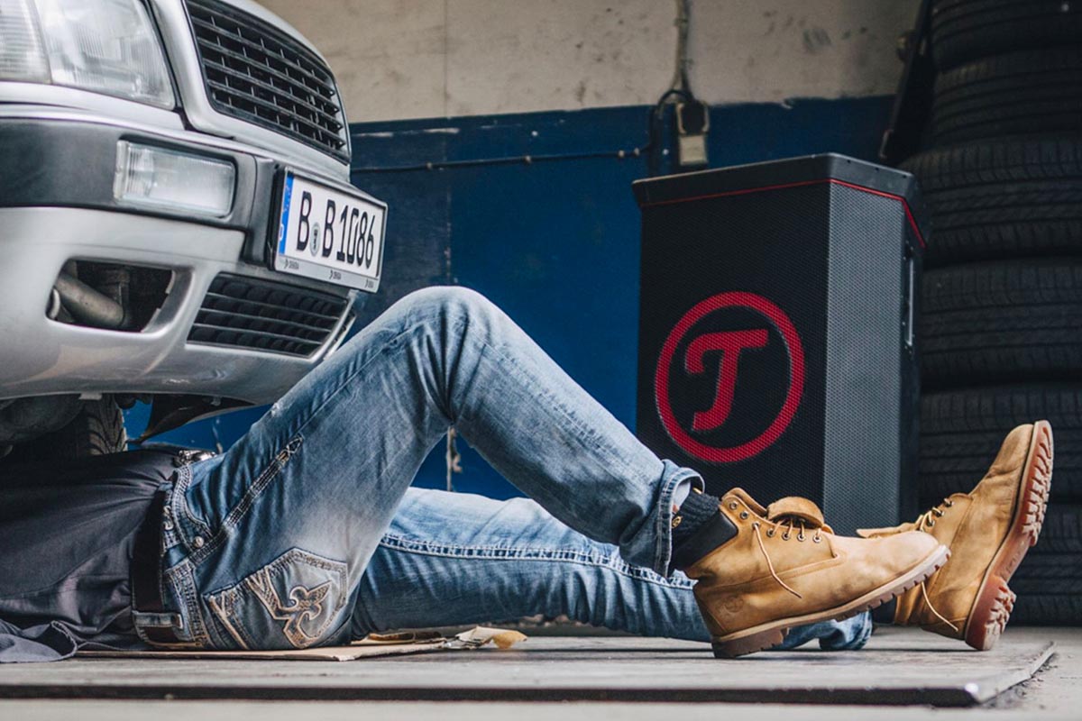 Ein Mann liegt auf dem Boden, um ein Auto von unten zu reparieren. Dahinter steht der Teufel Rockster Lautsprecher.