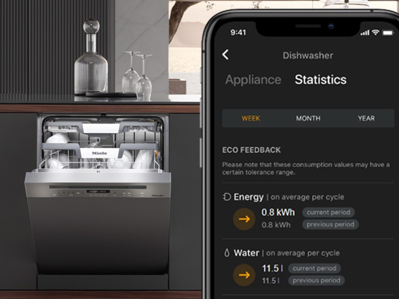 Ein Handy mit der Miele@home App ist zu sehen. Strom- und Wasserverbrauch werden angezeigt. Im Hintergrund ist ein Geschirrspüler.