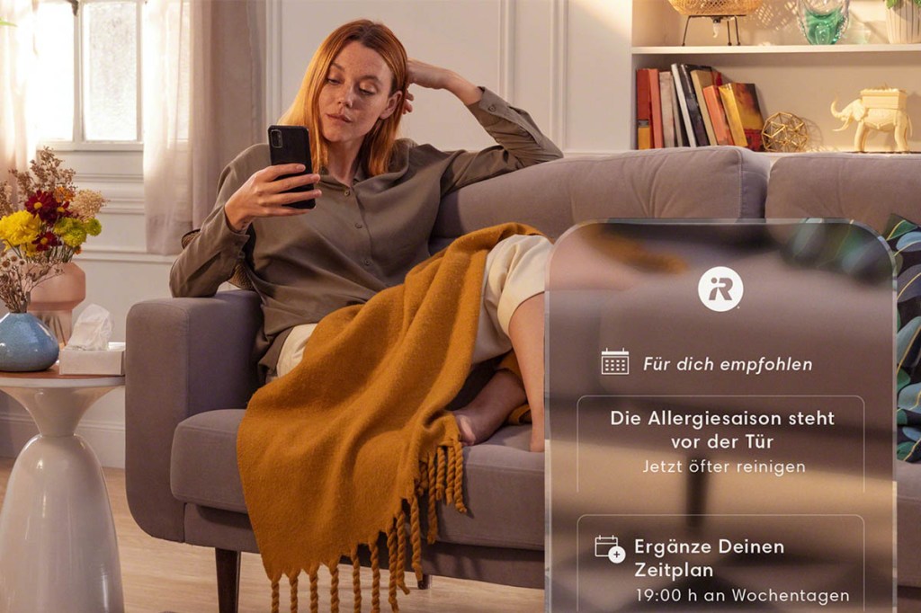 Eine Frau sitzt auf dem Sofa und schaut aufs Smartphone. Vergrößert ist das Display zu sehen mit Empfehlungen der iRobot Home App.
