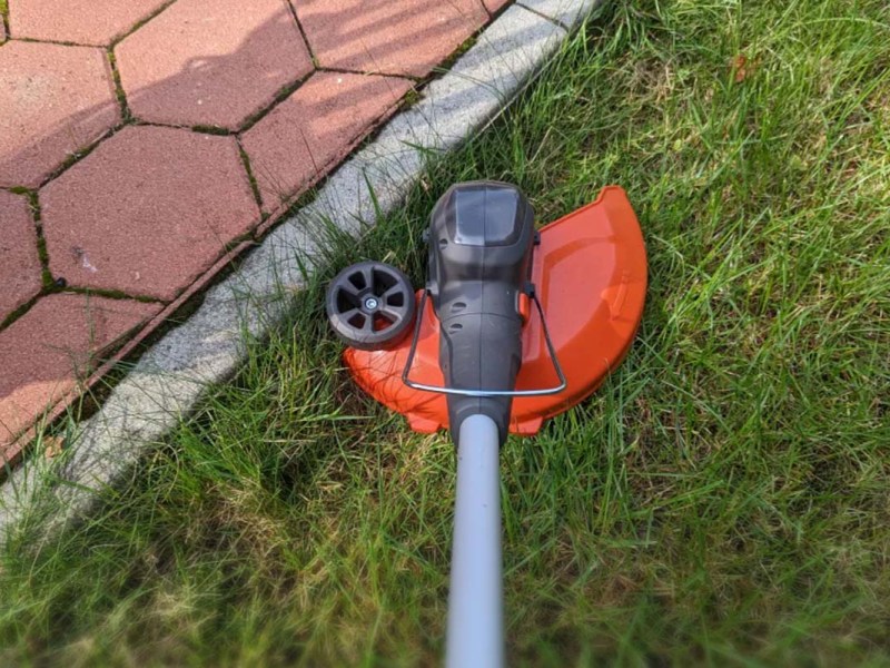 Anschnitt eines Rasentrimmers von oben, man sieht den Mähkopf, wie er an einer Rasenkante entlang eines Steinwegs mäht