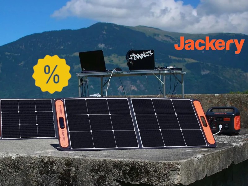 Aufgestellte Solarpanel vor Powerstation und Tisch mit Computer auf steinplatte vor Bergkulisse mit gelben Prozentzeichen links und Jackery Logo rechts